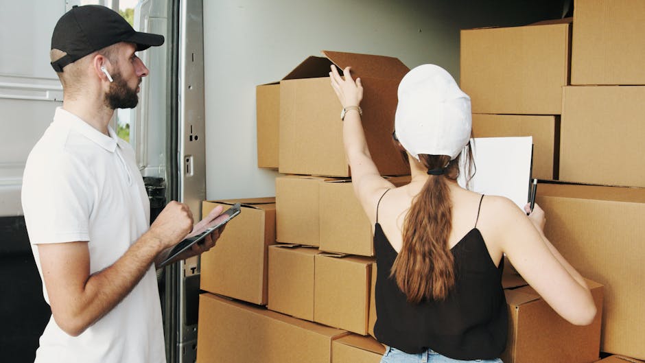 A man wearing a black cap, white t-shirt, and white earbuds is inspecting or taking notes on a clipboard while standing beside a partially opened delivery van. Inside the van, there are multiple stack