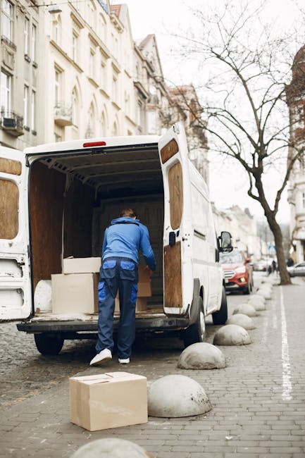 A man wearing a black cap, white t-shirt, and white earbuds is inspecting or taking notes on a clipboard while standing beside a partially opened delivery van. Inside the van, there are multiple stack