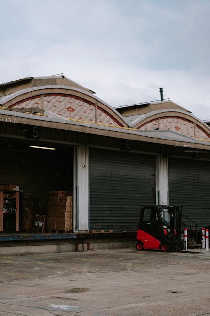 A warehouse with two large roller shutters, numbered 5 and 6, partially open revealing stacked cardboard boxes and packing materials inside. In front of the shutters, a red and black electric forklift is parked on an outdoor concrete loading area. The building has a curved roof with decorative brickwork, and the sky overhead is partly cloudy. The scene depicts a typical loading and unloading environment relevant to house removals and furniture transport services provided by Man with Van Blackwall, potentially during a home relocation process near East India Dock Road, with a focus on safety and efficient logistics for avoiding loading bay fines.