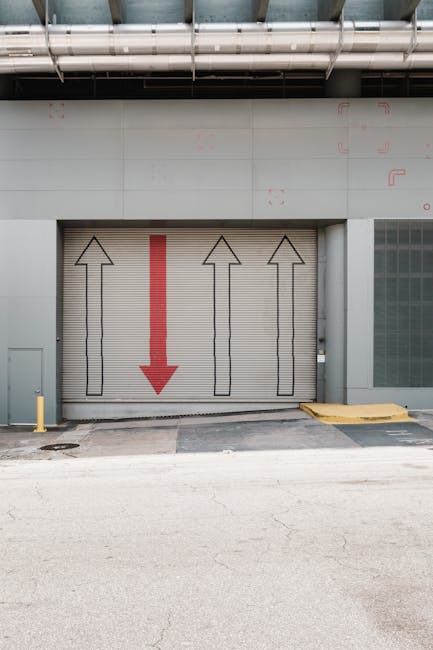A warehouse with two large roller shutters, numbered 5 and 6, partially open revealing stacked cardboard boxes and packing materials inside. In front of the shutters, a red and black electric forklift is parked on an outdoor concrete loading area. The building has a curved roof with decorative brickwork, and the sky overhead is partly cloudy. The scene depicts a typical loading and unloading environment relevant to house removals and furniture transport services provided by Man with Van Blackwall, potentially during a home relocation process near East India Dock Road, with a focus on safety and efficient logistics for avoiding loading bay fines.