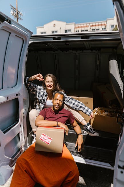 A man and a woman are standing beside an open moving van filled with stacked cardboard boxes in various sizes, some wrapped in plastic. The man, wearing a black cap, white t-shirt, and white wireless 