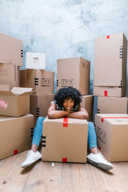 A young woman with curly black hair, wearing a light grey t-shirt, blue jeans, and white sneakers, sits on a light wooden floor inside a room filled with numerous cardboard boxes used for packing duri
