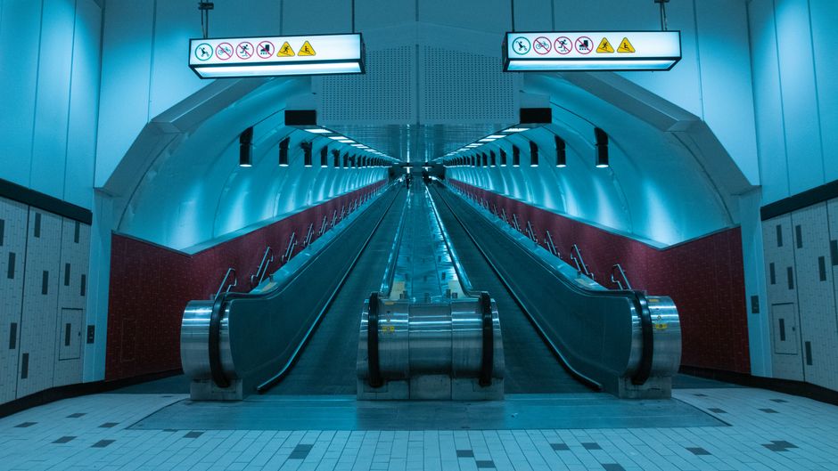 A long, illuminated underground tunnel at Blackwall with curved metallic walls and ceiling panels, featuring several evenly spaced fluorescent light fixtures. The perspective shows the moving walkway 