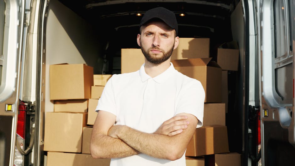 A man wearing a black cap and a white polo shirt is standing indoors, holding a clipboard and pen while examining a large collection of cardboard boxes stacked against a wall. The boxes are of various