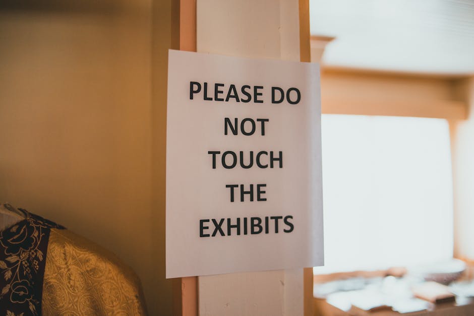 A white paper sign with bold black text reading 'PLEASE DO NOT TOUCH THE EXHIBITS' is attached to a vertical wooden post inside a premises, likely a house or storage area. The sign is positioned near a doorway or entrance, with part of a patterned fabric or upholstery visible on the left side of the image. The background shows a well-lit interior space with a large window or opening allowing natural light to illuminate the room. The scene suggests that this area could be part of a property scheduled for a home relocation or furniture transport process, which is handled by a professional removals service such as Man with Van Blackwall. The setting indicates a controlled environment where delicate or valuable items are protected from unnecessary handling during packing, moving, or loading activities, which are common steps in moving and clearance projects.