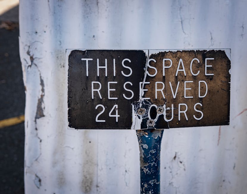 A close-up image of a weathered metal parking sign mounted on a blue metal pole, which is fixated to a wall. The sign reads 'THIS SPACE RESERVED 24 HOURS' in white uppercase letters on a black background, although the sign shows signs of rust, peeling paint, and damage, with chipped edges and some rust spots. The wall behind the sign is white with visible cracks and weathering. In the background, part of a concrete or asphalt surface is visible, suggesting an outdoor setting. This scene relates to urban environments where house removals or moving services by Man with Van Blackwall often require parking permits or reserved spaces during the loading and unloading process of furniture and boxes for home relocation or furniture transport purposes.