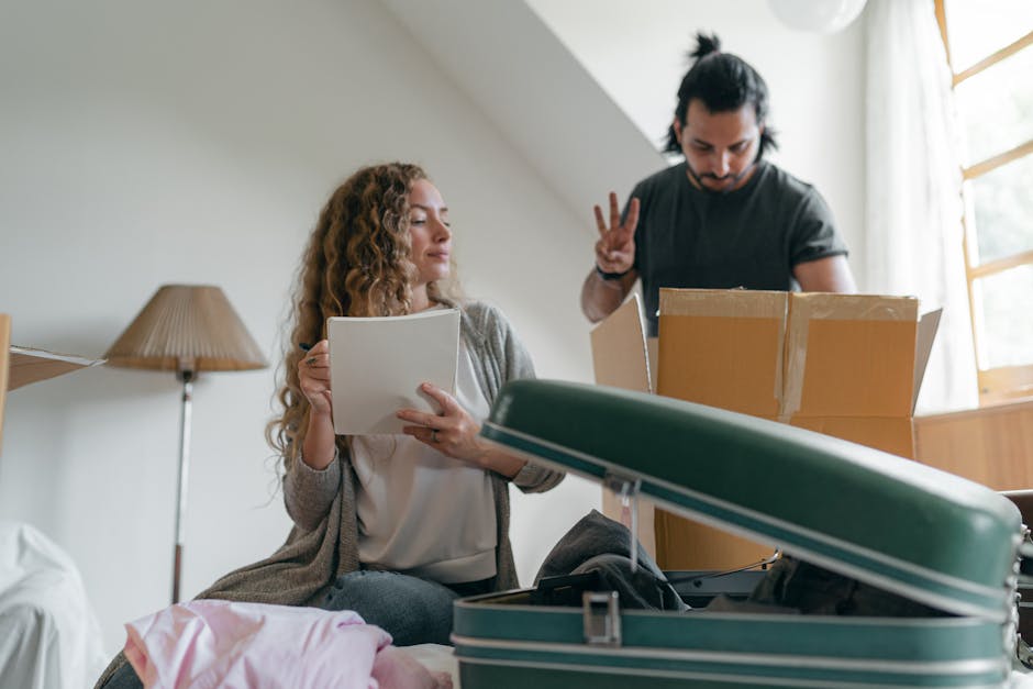A woman with long, curly hair and light skin is sitting on a bed inside a room, holding a clipboard or a packing list and observing a man with dark, tied-back hair and light skin who is standing next to a cardboard box on a table. The man is making a peace sign with his right hand, both of them surrounded by various moving supplies including a partially open, teal-colored hard-shell suitcase in the foreground and several cardboard boxes in the background. The room is naturally lit through a window with beige curtains pulled to the side, and a wooden bedside lamp with a woven shade is visible on a side table. This scene captures a home relocation process involving packing materials, luggage, and planning, relevant to house removals and furniture transport, with the activities taking place in a domestic interior environment as part of a professional move managed by Man with Van Blackwall.