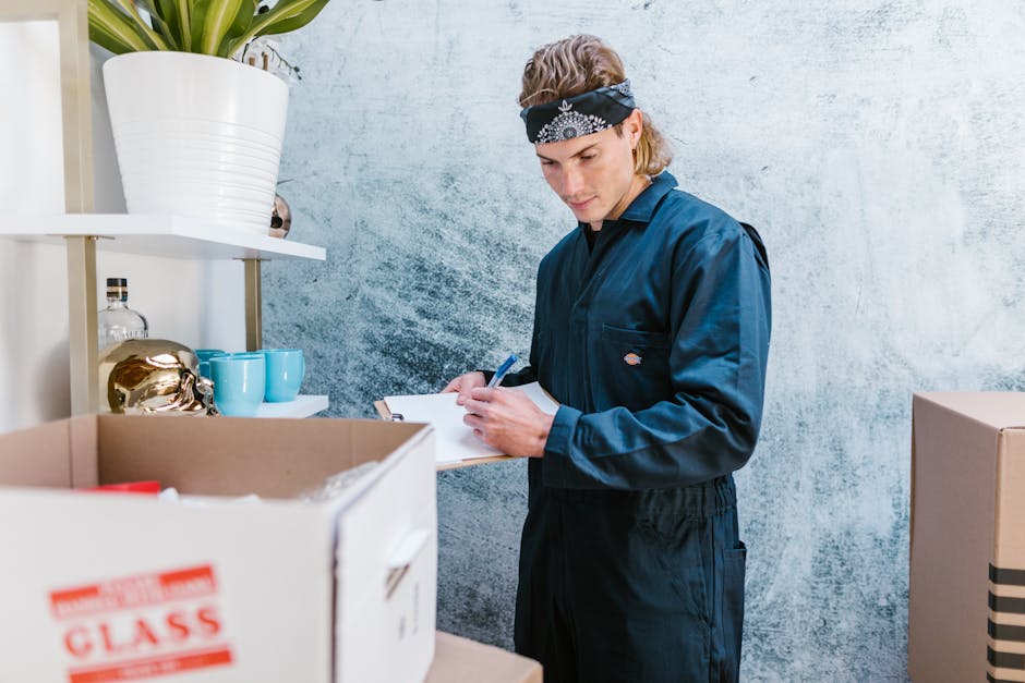 A man wearing a dark blue uniform and a black bandana with white patterns is standing in a room with a textured light grey wall, holding a clipboard and a pen while inspecting or taking notes during a home relocation process. In front of him, there are open cardboard boxes containing various household items, including a red item, and packaging materials. To his right, a white shelving unit holds decorative objects such as a gold-colored sculpture, two turquoise cups, and a large white ceramic vase with a plant. The scene is lit with natural or soft artificial light, and the presence of packed boxes suggests that the individual is involved in coordinating or overseeing the furniture transport and packing procedures as part of the house removals service offered by Man with Van Blackwall, supporting efficient logistics for moving into E14 (Blackwall) flats.