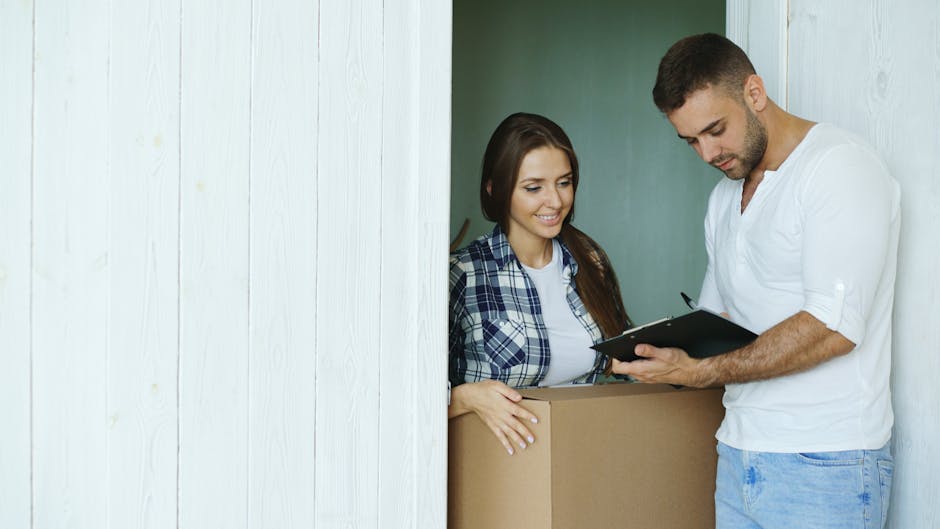 A woman with long, curly hair and light skin is sitting on a bed inside a room, holding a clipboard or a packing list and observing a man with dark, tied-back hair and light skin who is standing next to a cardboard box on a table. The man is making a peace sign with his right hand, both of them surrounded by various moving supplies including a partially open, teal-colored hard-shell suitcase in the foreground and several cardboard boxes in the background. The room is naturally lit through a window with beige curtains pulled to the side, and a wooden bedside lamp with a woven shade is visible on a side table. This scene captures a home relocation process involving packing materials, luggage, and planning, relevant to house removals and furniture transport, with the activities taking place in a domestic interior environment as part of a professional move managed by Man with Van Blackwall.