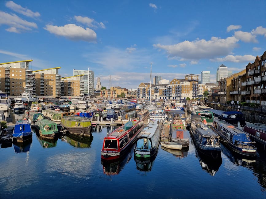 A view of a marina with multiple boats moored along the dock, including narrowboats and small leisure vessels, some covered with tarps or fitted with protective sheeting. Behind the marina, there are modern residential buildings with balconies and large windows, contrasting with older brick structures. The sky is partly cloudy with a bright blue background, and a few high-rise buildings can be seen in the distant skyline. The scene captures a typical urban riverside environment in London, relevant to home relocation and furniture transport services offered by Man with Van Blackwall during moving processes from East India Dock Rd to Canary Wharf, with boats and buildings reflecting the logistics involved in house removals in this area.