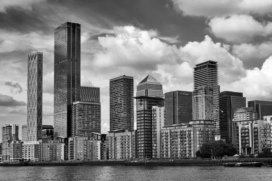 Black and white photograph of a city skyline featuring tall modern skyscrapers with varying architectural styles along the waterfront, with some buildings displaying glass facades and others with concrete exteriors. The scene captures a partly cloudy sky, with large clouds casting shadows over the buildings. In the foreground, there are mid-rise residential buildings with balconies, situated close to the water’s edge. The image depicts a dense urban environment typical of a central business district, highlighting the contrast between residential and commercial structures, which is relevant for home relocation and furniture transport services provided by Man with Van Blackwall during house removals or moving processes from East India Dock Road to Canary Wharf.