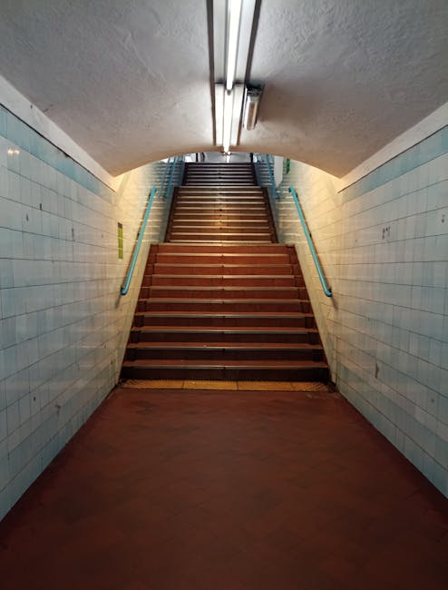 Photograph of a narrow underground staircase with wooden steps leading upward, enclosed by tiled walls painted in light blue. A handrail is attached to each side of the staircase, painted in a contrasting darker blue. The ceiling above the stairs has a curved, tunnel-like shape with fluorescent lighting mounted along its length, illuminating the area evenly. The scene appears to be in a public transportation or pedestrian underpass area, with no furniture or packing materials visible. This setting is relevant to home relocations, as its stairs and access points could relate to the logistics of moving belongings in and out of properties, with the image possibly illustrating a typical route for furniture transport during an underground corridor move managed by [COMPANY_NAME], supporting efficient packing and loading processes for a house removal or flat relocation.