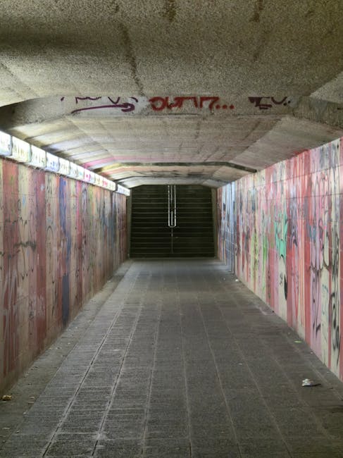 A close-up view of a steep metal staircase with yellow safety handrails on both sides, leading upward inside a building, with the steps made of black non-slip material and yellow edging. Several steps include white stickers with black text reading 'PLEASE KEEP LEFT,' indicating a directional flow for pedestrian movement. The staircase is surrounded by an industrial environment with visible beams and structural elements overhead, and natural light filtering into the space. This staircase is part of a setting where house removals or moving services, such as those provided by Man with Van Blackwall, may typically be involved in facilitating home relocation or furniture transport through building access points.