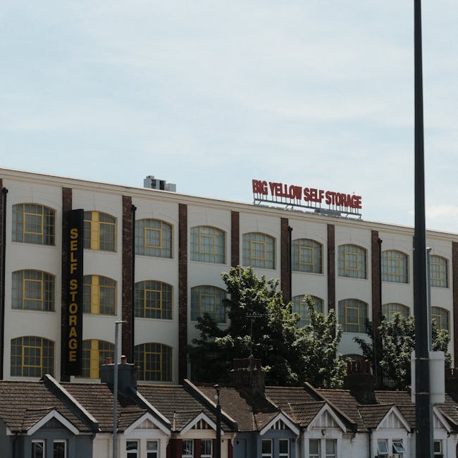 A large multi-story building with a white facade and brick accents, featuring numerous tall, arched windows with yellow frames. Prominent black vertical signage on the left side reads 'SELF STORAGE,' indicating a self-storage facility. On the roof, a red sign displays 'BIG YELLOW SELF STORAGE' in capital letters. In front of the building, there are small residential-style houses with pitched roofs and chimneys, alongside several trees with green foliage partially obscuring the lower part of the building. The sky above is partly cloudy with patches of sunlight. The image appears to be taken during daytime, possibly in an urban area where a moving or packing process might be taking place, consistent with services provided by companies like Man with Van Blackwall during home relocation or furniture transport.