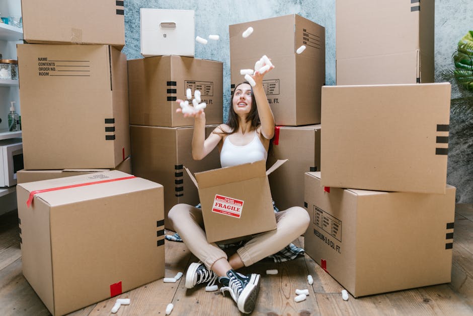 A young woman sits on a wooden floor surrounded by numerous large cardboard moving boxes, some sealed with red or black tape, with a few labeled with contents and room information. She is inside a room with plain light blue walls and a potted plant visible on the right side. The woman is smiling and looking up while tossing packing peanuts into the air, with some falling back around her. She wears a white sleeveless top, beige pants, and black and white sneakers, and appears to be enjoying the packing or unpacking process during her home relocation. Behind her, at least one of the boxes is partially inside an open cardboard box, and additional boxes are stacked against the walls, suggesting an organized packing scene. The setting includes a small white cupboard or shelf on the left, and the scene conveys a domestic environment associated with furniture transport, packing materials, and moving activities, aligned with house removals and local moving services by Man with Van Blackwall.