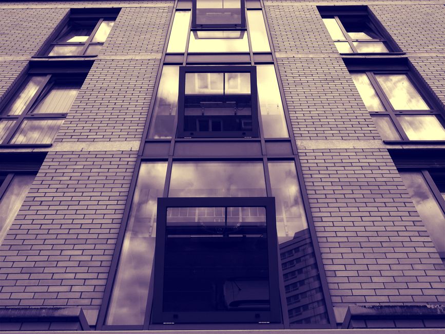 Photograph of the exterior of a multi-storey residential building with a modern brick facade and large, reflective glass windows. The image is taken from a low angle, looking up at the building, showing several windows on each floor. Some windows have partially visible curtains or blinds, and reflections of nearby structures and the cloudy sky are visible in the glass panes. The building is situated on a pavement, with no vehicles or moving equipment in sight. The image provides a clear view of the building's architectural features, highlighting its height and contemporary design, which may present challenges for home relocation or furniture transport, particularly in tight spaces such as narrow stairwells or small entrances. Man with Van Blackwall may assist with moving projects involving similar flats by offering professional removals and packing services to overcome such logistical challenges.