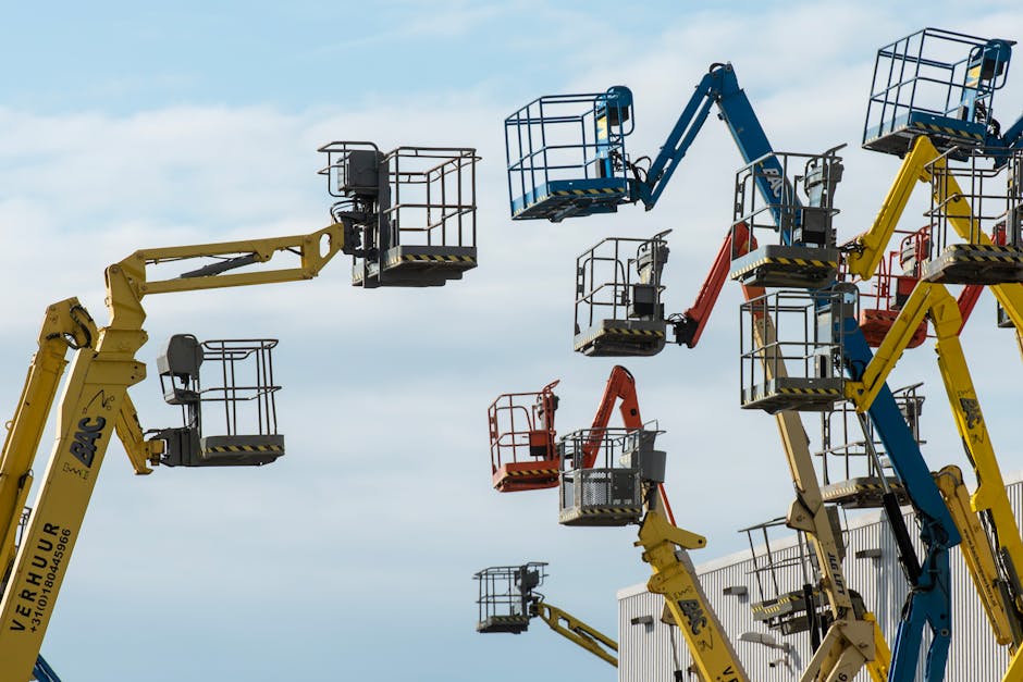 Multiple articulated boom lifts in yellow, blue, and red, positioned outdoors on a paved surface, with some elevated and others lowered, supporting work at various heights against a cloudy sky. The lifts have metal baskets with safety railings and extend upward, appearing to be used for maintenance or construction activities near a white building. The scene exhibits a variety of equipment including hydraulic arms, control panels, and safety markings, with some lifts tipped slightly at different angles. The environment reflects professional height access in a commercial or residential setting, consistent with tasks related to home relocation or property improvements. Man with Van Blackwall occasionally visible in context as part of the logistics for moving or site clearance services.