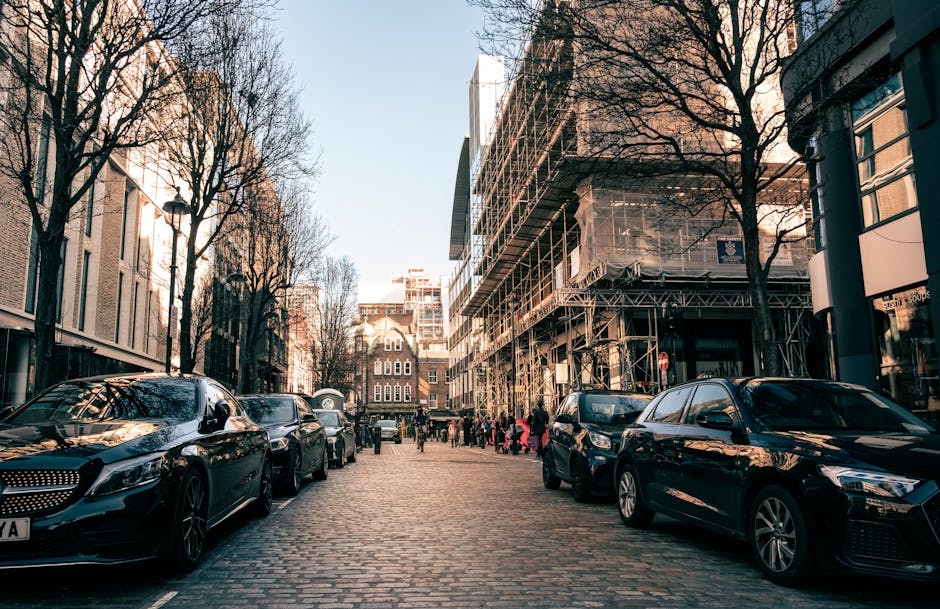 A street scene in Tower Hamlets showing parked cars along a cobblestone road with a construction site on the right side, partially covered with scaffolding and protective sheeting. On the left, leafless trees line the street, with buildings and a lamppost visible in the background. Pedestrians can be seen walking and standing near the sidewalk. The image is taken during daylight with clear weather, highlighting the urban environment typical of Blackwall in London. This setting provides context for home relocation or furniture transport activities, as seen in professional removals services such as Man with Van Blackwall, who manage loading and moving processes in such urban areas.