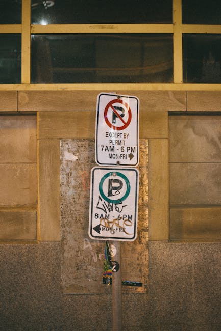 Close-up of a metal parking restriction sign mounted on a wall, showing two sections: the top indicating no parking except by permit from 7 am to 6 pm, Monday to Friday, and the bottom indicating permit parking from 8 am to 6 pm, Monday to Friday. The sign is attached to a weathered metal pole, which is secured to a stone wall beneath a large window with a yellow frame. The wall behind the sign appears to be composed of stone or brick with a smooth finish, and the scene is illuminated by artificial lighting, suggesting an indoor or shaded area. This setting may be relevant to the process of house removals or furniture transport, where parking restrictions impact loading and unloading activities. Man with Van Blackwall offers services in such urban environments within Tower Hamlets, assisting with packing, home relocation, and transportation coordination.
