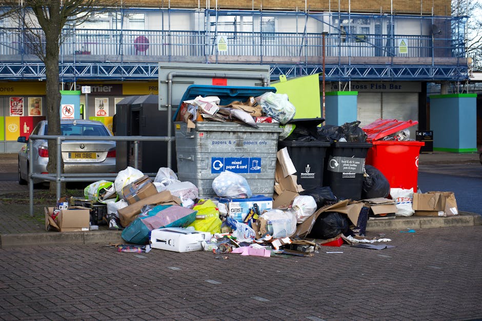 An overflowing grey mixed paper and card recycling bin situated on a pavement in front of a commercial building, with its lid open and various items such as crumpled paper, cardboard boxes, plastic bags, and packaging materials spilling onto the surrounding area. To the left of the recycling container, there are additional black and red waste bags and flattened cardboard boxes. Behind the bin, part of a parked silver car is visible, along with metal railings separating the vehicle from the waste area. The background features a building with storefronts, including signage for a fish bar and other shops, and a second-floor construction scaffold covered with blue netting. The scene captures a typical urban environment where waste collection and recycling are part of residential or commercial property management, relating to house removal or moving services provided by Man with Van Blackwall for efficient furniture transport and packing support.