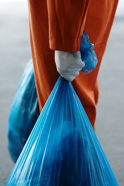 A person wearing an orange high-visibility jacket and white gloves is holding a large blue plastic garbage bag filled with waste, standing outdoors on a paved surface. The background is blurred, suggesting an open, urban environment. This image relates to waste disposal and clearance services often involved in house removals or property cleanouts, which are part of comprehensive relocation processes handled by companies like Man with Van Blackwall. The scene captures the moment of waste collection or disposal, related to managing bulky waste during home relocation or removal projects, emphasizing the importance of proper disposal before or after moving activities. The packaging material and handling equipment indicate a professional approach to waste management, supporting efficient clearance and preparation of a property for a move.