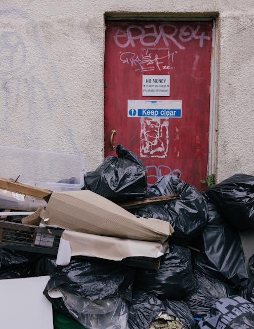 An overflowing grey mixed paper and card recycling bin situated on a pavement in front of a commercial building, with its lid open and various items such as crumpled paper, cardboard boxes, plastic bags, and packaging materials spilling onto the surrounding area. To the left of the recycling container, there are additional black and red waste bags and flattened cardboard boxes. Behind the bin, part of a parked silver car is visible, along with metal railings separating the vehicle from the waste area. The background features a building with storefronts, including signage for a fish bar and other shops, and a second-floor construction scaffold covered with blue netting. The scene captures a typical urban environment where waste collection and recycling are part of residential or commercial property management, relating to house removal or moving services provided by Man with Van Blackwall for efficient furniture transport and packing support.